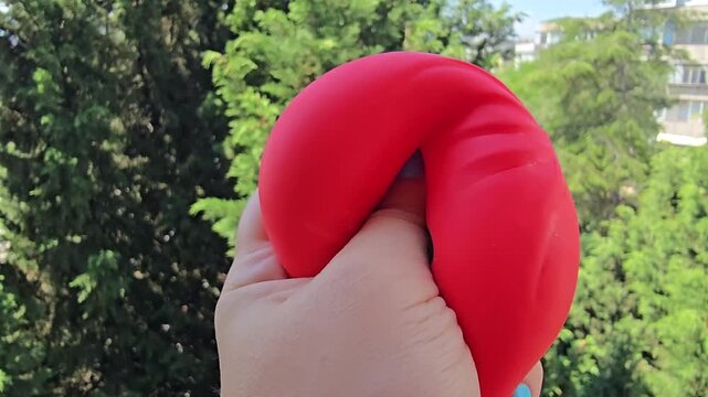 Red antistress bao toy with kawaii smiley face held in hand outdoors against green background, glossy squishy stress relief item