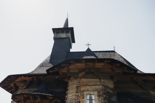 Old wooden church with steep shingled roof. Stone base supports intricate wooden beams. Cross adorns peak beneath cloudy sky. Vantage view highlights rustic architectural detail