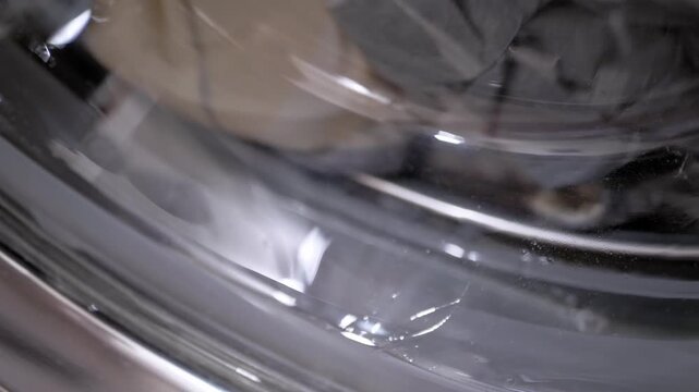 A close-up view of clothes tumbling in soapy water inside the rotating drum of a front-loading washing machine. Concept of household chores, domestic hygiene, and daily appliance use.