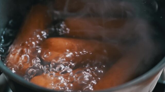 Thick brown sausages boil vigorously inside a dark pot of bubbling water with rising steam. Concept of fast food preparation, quick home cooking, and unhealthy dietary eating habits.