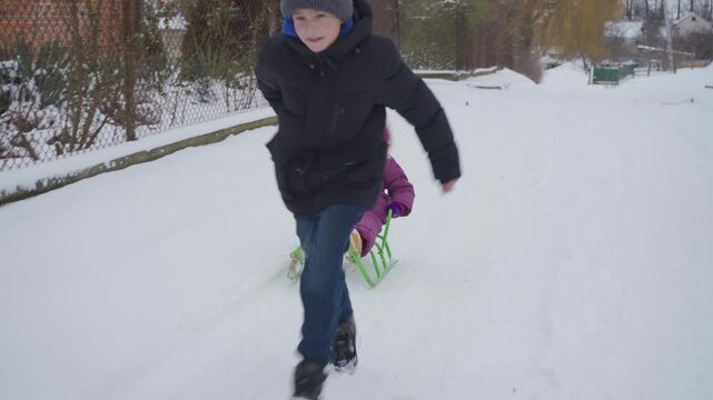 Older Brother Pulling Sled with Little Sister on Snowy Road, Boy Running and Sledding Cute Toddler Girl in Winter 