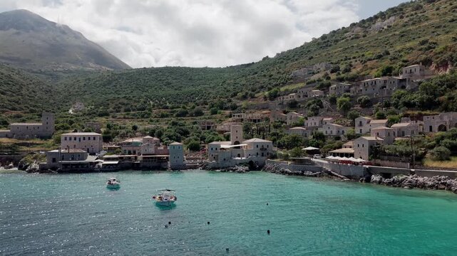 Drone shot of Limeni village traditional stone tower houses to rocky coastline on turquoise waters, fishing boats with lush green steep mountains in the backdrop under cloudy sky