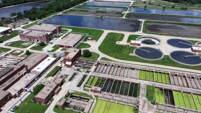 Forward drone shot above large sludge settling ponds at a water reclamation facility with excavator visible and brick operations buildings in foreground.