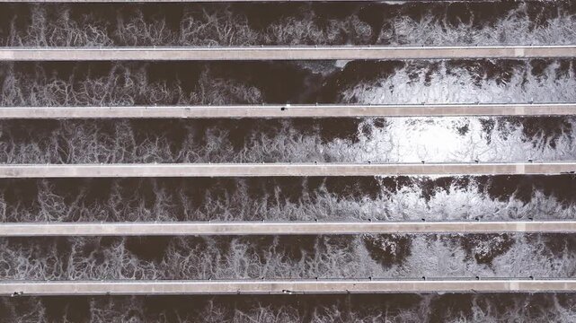 Top down drone shot descending above active aeration channels at a wastewater treatment facility revealing churning water and foam patterns.