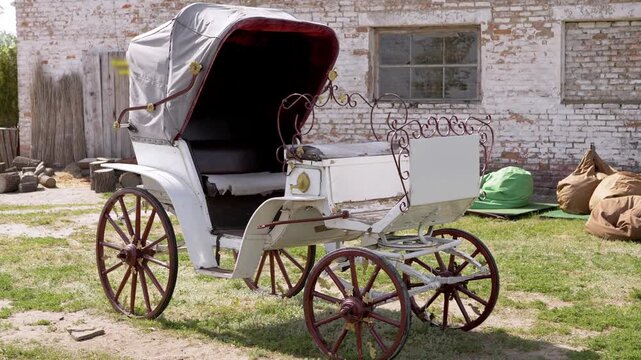 A vintage white horse carriage with burgundy wheels sits parked on grass near a brick wall. Concept of historical transportation, romantic weddings, and nostalgic classic travel.