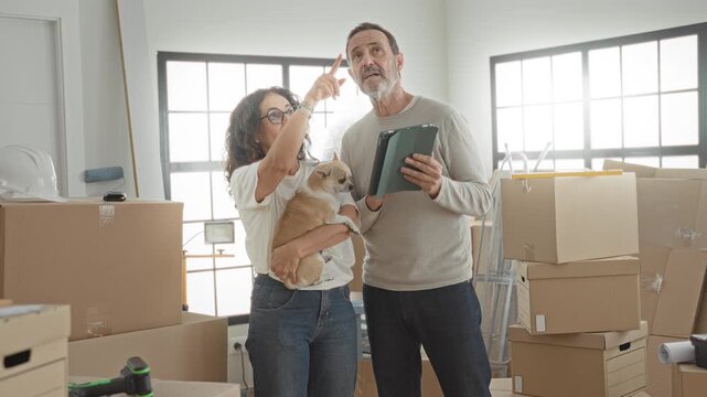 Man and woman carry boxes and hold a small dog while woman points and man reads a tablet in a building; hopeful planning.