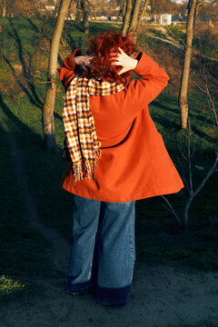 Person in orange coat standing outdoors adjusting hair autumn park scene