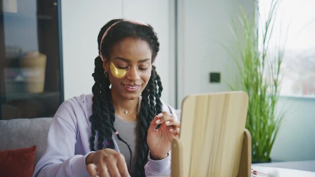 A woman is applying a gold face mask while sitting at a table. She looks at her reflection in a mirror. The room is bright and contains skincare products.
