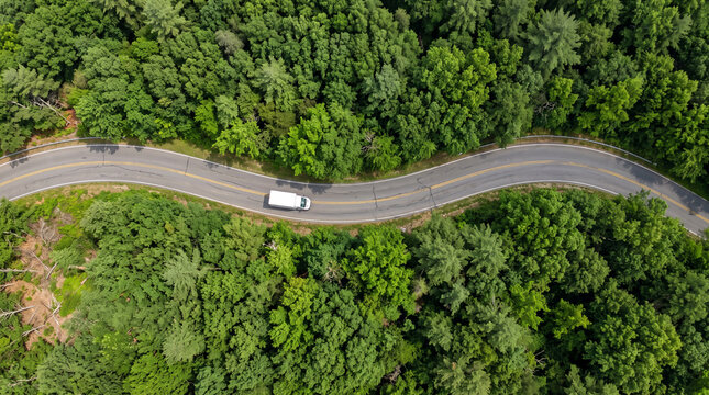 Aerial view of a white delivery truck driving on a winding asphalt road through a dense green forest during summer