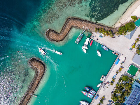 Aerial drone view of Maafushi island jetty with tour boats speeding through the entry between breakwaters. Nautical vessel transportation system for Maldives travel industry