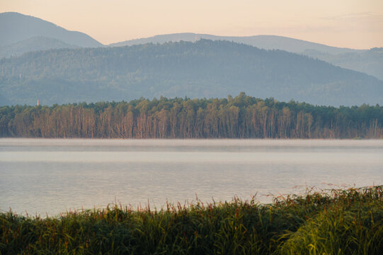 Distant view of mist rising above a calm lake with forested hills and mountains in warm morning light.