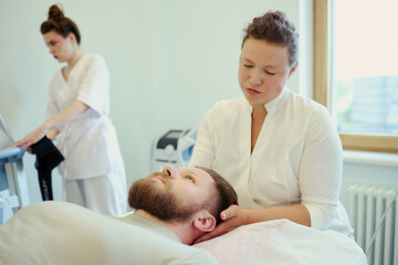 Fototapeta premium Young adult Caucasian woman therapist massaging neck of bearded Caucasian man during rehabilitation session while female colleague checking monitor. For wellness clinic marketing