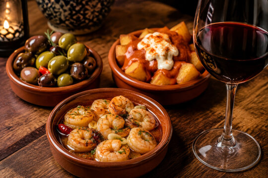 Glass of red wine with Spanish tapas dishes including garlic prawns, patatas bravas and olives. Traditional Mediterranean dinner culinary concept of assorted appetizer food plate.