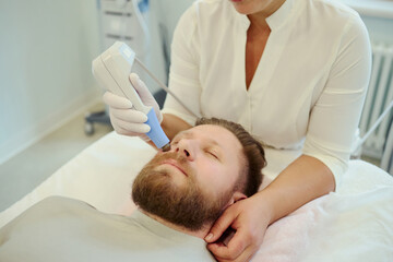 Fototapeta premium Close up of Caucasian young adult man receiving facial skin treatment from female clinician, targeting beard area for grooming and aesthetic care. Ideal for mens skincare marketing