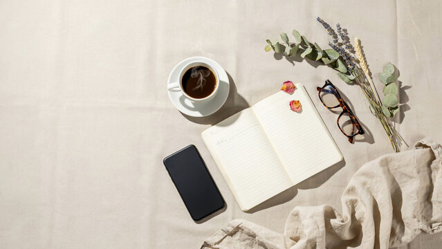 A serene workspace setup with a cup of coffee, notebook, phone, glasses, and flowers on a tablecloth.