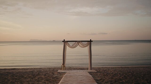 A simple and elegant wooden wedding arch draped with white fabric on a sandy beach, overlooking the vast ocean at calm sunset, ideal for a destination wedding.