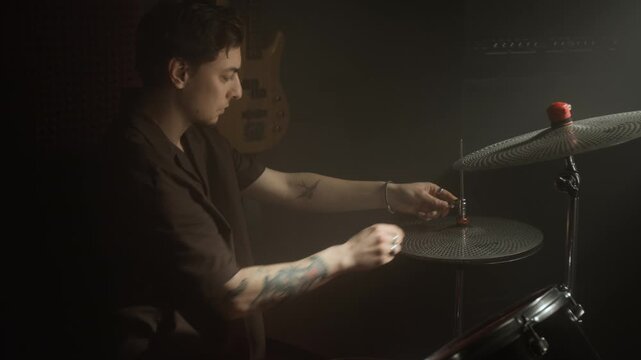 Young male drummer with tattoos adjusting hi-hat cymbal on drum kit in dark studio and picking up drumsticks. Footage for music school advertising or instrument promos