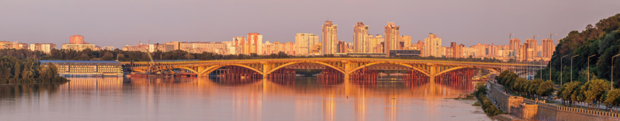 Fototapeta premium Kyiv, Ukraine - July 2 2026: View of Metro Bridge across the Dnipro River in Kyiv, Ukraine, glowing in warm evening light with reflections on calm water and the modern city skyline in the background.