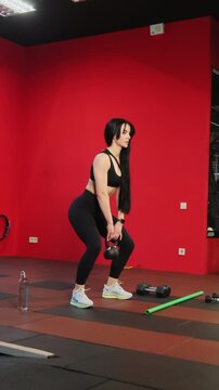 Woman performs kettlebell squat against red wall in gym zone. Athlete lowers hips with steady control keeping chest lifted through strong movement