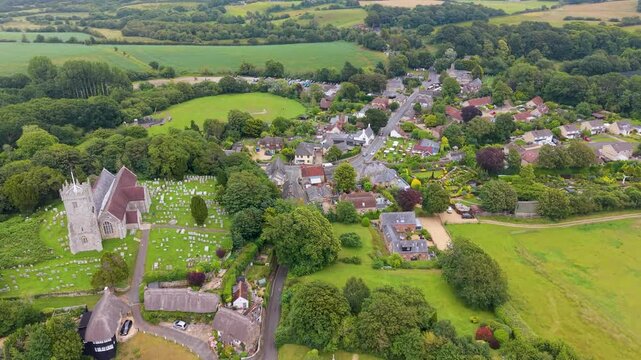 Godshill, Isle of Wight drone flight over All Saints Church and village rooftops, passing the Model Village attraction and Central Mead park, with green fields and wooded countryside.
