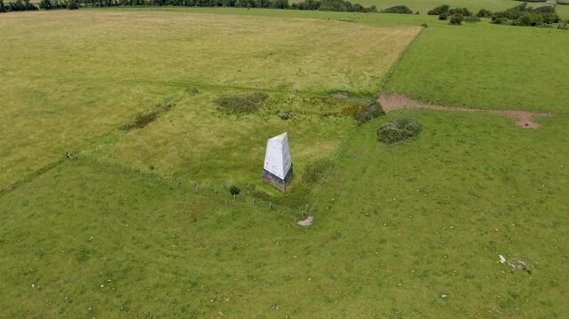 Ashey Down sea mark obelisk as an ascending drone shot holds focus on the landmark, creating parallax as patchwork farmland and hedgerows shift behind on the Isle of Wight, England.