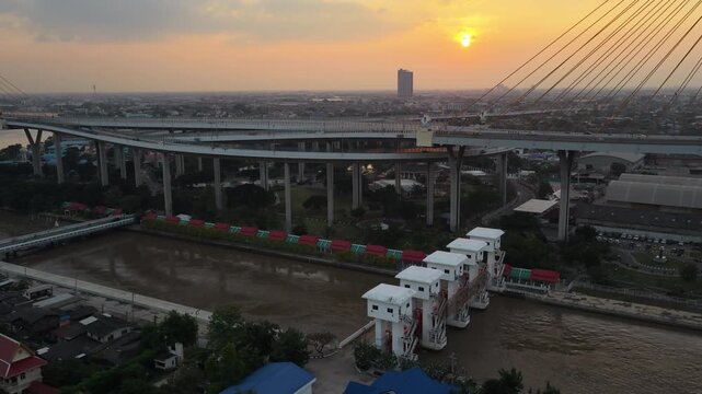 Bhumibol Bridge interchange above canal floodgates during sunset in Bangkok, drone medium overview tilt up from water to city