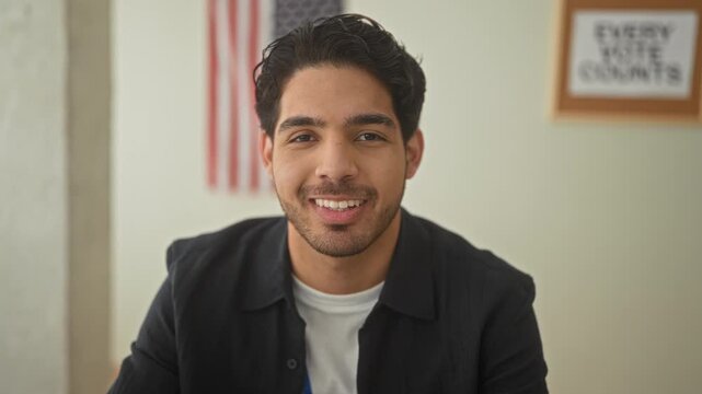 Man smiling showing teeth and looking at camera beside an american flag and every vote counts poster in a polling building; civic pride hopeful.