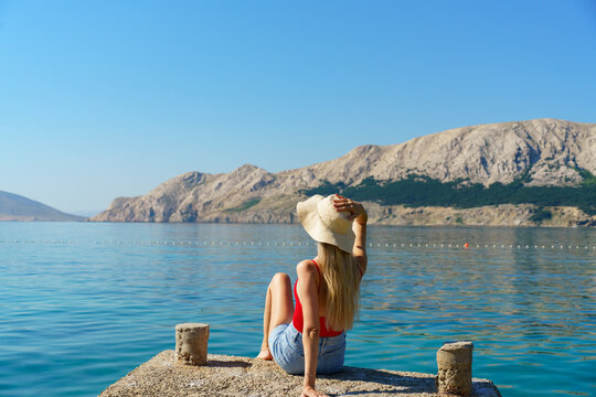 Young blonde woman sitting on a seaside pier holding her sun hat while enjoying the panoramic view of the calm Adriatic Sea and rocky mountains in Ba&scaron;ka, Krk Island, Croatia