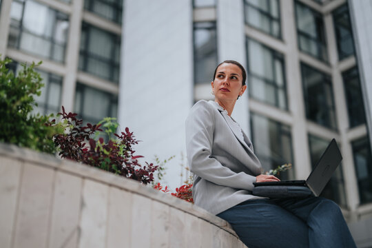 A businesswoman in a gray suit works on a laptop while seated on a planter ledge. She uses her computer in an outdoor corporate plaza with high-rise offices in the background.