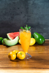 Vibrant Citrus Elixir: A refreshing glass of orange juice, adorned with a sprig of mint, sits amidst a colorful medley of fresh fruits, including watermelon, melon, avocado, and oranges. © andreas