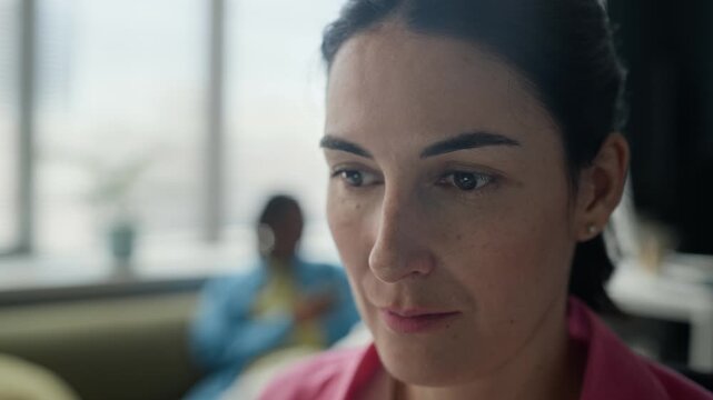 Closeup of young adult Caucasian woman putting on headphones and closing eyes to relax in office, colleague working in background. Perfect for corporate wellness and mental health at work videos