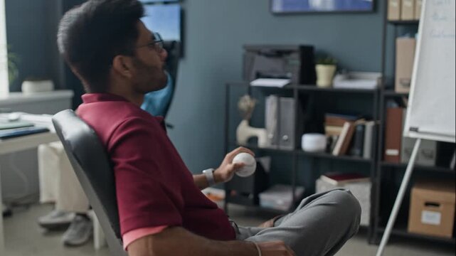 Young adult Indian man sitting in office chair, tossing baseball and looking at flipchart. Footage is perfect for corporate environment and showcasing creative workplace culture