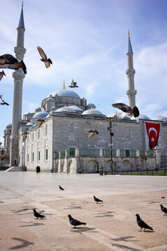 A flock of pigeons takes flight in front of the New Mosque with twin tall minarets and a Turkish flag in an open plaza in Istanbul.