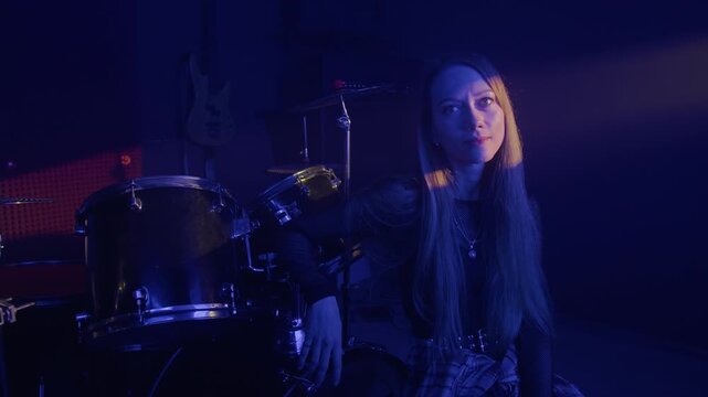 Portrait of young Caucasian woman sitting next to drum kit in music studio, turning head to look at camera. Footage for music school advertising or band recruitment campaigns