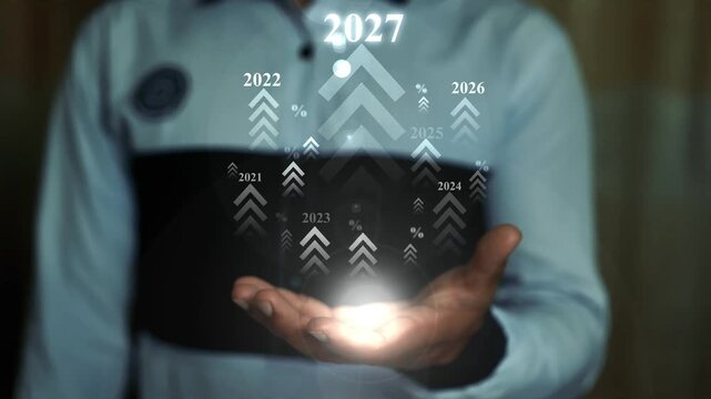Businessman in a futuristic outfit holds a glowing light hologram in an open hand with 2027 animation on the screen, Holding glowing technology innovation and a forward-looking vision in a dark settin
