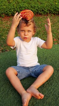 Baby boy sits on grass, balancing large croissant on his head in playful trick. In slow motion, he lowers his hands and concentrates, but croissant slips and falls. Candid outdoor moment