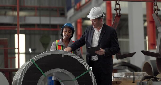 A senior manager and an African female worker in a safety vest use a digital tablet to inspect steel coils stored in a warehouse under an overhead crane hook discussing stock quality