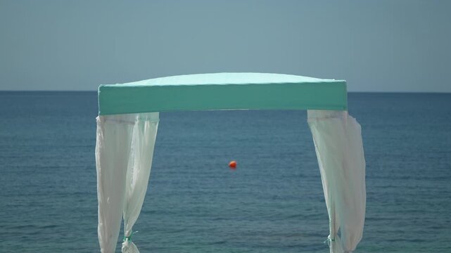 Beach wedding canopy stands centered on a sandy shore under clear blue skies. Static wide shot.