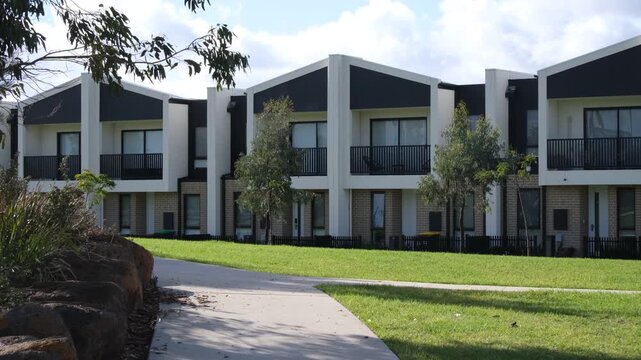 Modern two storey townhouses with dark roofline and balconies overlooking a footpath and lawn in an public green space in outer suburban Melbourne Australia. Contemporary medium density housing