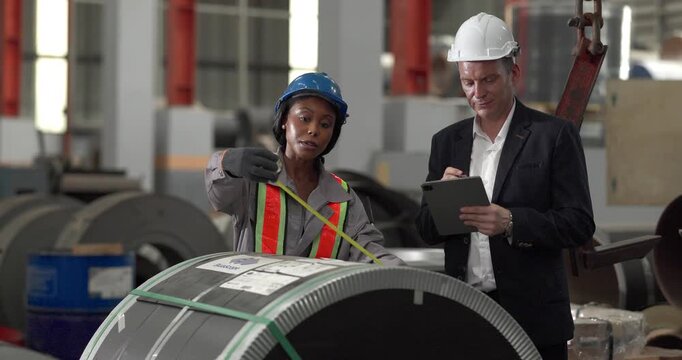 A senior manager and an African female worker in a safety vest use a digital tablet to inspect steel coils stored in a warehouse under an overhead crane hook discussing stock quality