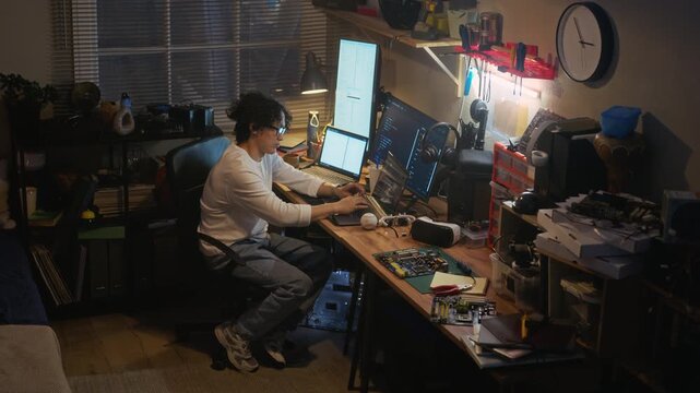 Asian young adult man typing on laptop at cluttered desk with multiple monitors in home office. Ideal for tech startup marketing, software development, or remote IT work concepts