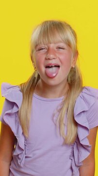 Cheerful funny young blonde school lady showing tongue making faces at camera, fooling around, joking, aping with silly face, teasing, bullying, abuse. Preteen female child kid on yellow background