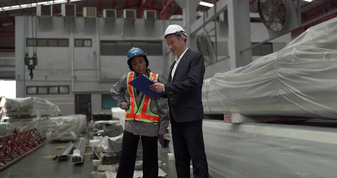 A senior manager and an African female worker in a safety vest use a digital tablet to inspect steel coils stored in a warehouse under an overhead crane hook discussing stock quality