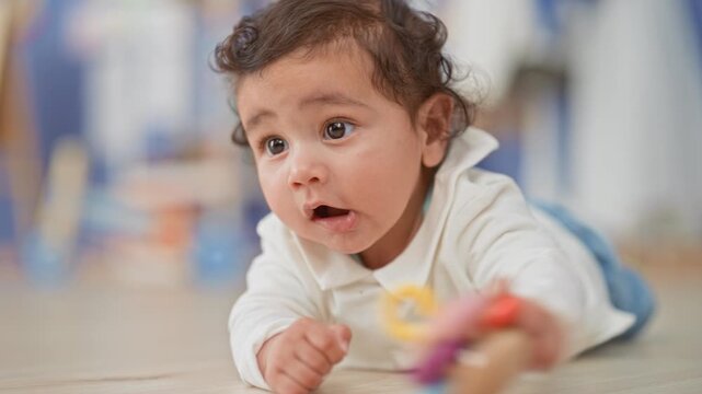 Toddler girl reaching with hand and holding a colorful rattle while lying on the floor in a studio; curious.