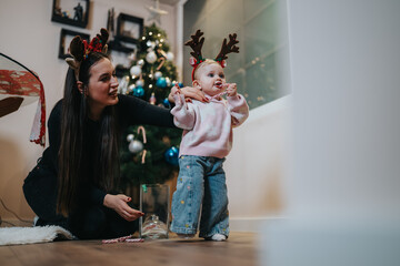 Fototapeta premium A mother helps her young child, both wearing holiday antler headbands, in a warm Christmas moment. They stand by a glowing tree inside a cozy home, creating a joyful family scene.