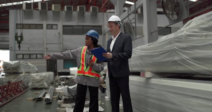 A senior manager and an African female worker in a safety vest use a digital tablet to inspect steel coils stored in a warehouse under an overhead crane hook discussing stock quality
