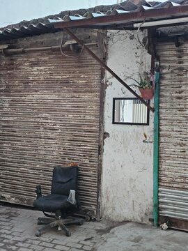 An abandoned street corner in Delhi features a worn-out office chair beside a weathered wall with sliding shutters, reflecting a sense of urban decay and solitude.