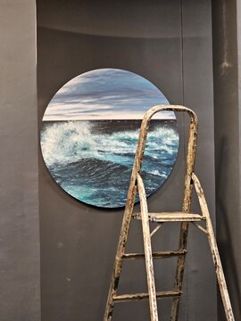 A circular painting of ocean waves is displayed on a gray wall, alongside a worn ladder, creating a serene yet artistic atmosphere in the setting.