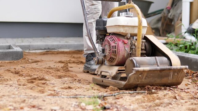 Low angle view shows worker legs beside plate compactor on sandy ground at residential jobsite, with curb, concrete edge, wheelbarrow, and machinery near building exterior in