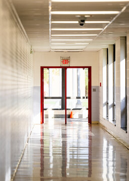 Empty school hallway with a glossy reflective floor leading toward red framed glass doors and a backlit exit sign. No people visible, well suited to safety, evacuation, and education concepts.